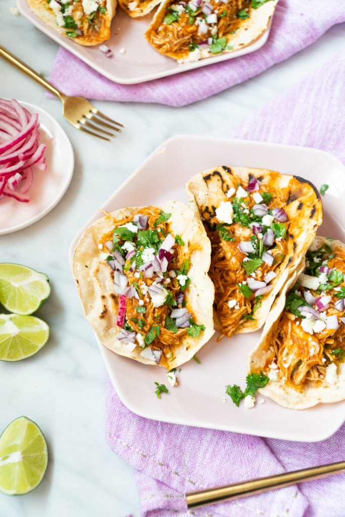 Chicken Tinga Tacos sprinkled with red onion, cilantro, and goat cheese with limes on the side