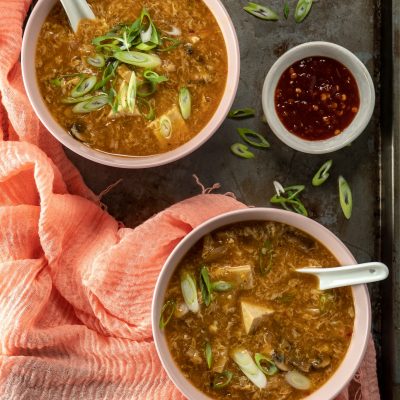 Bowls of warm Hot and Sour Soup sprinkled with green onions and served with a side of chili paste.